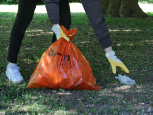 Eine Person mit einem orangefarbenen Müllsack und Handschuhen sammelt Müll auf einer Wiese.