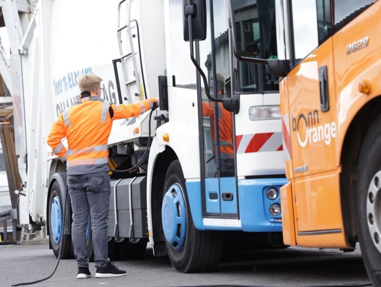 Ein Mitarbeiter der bonnorange lädt ein E-Abfallsammelfahrzeug neben einem orangenen Abfallsammelfahrzeug.