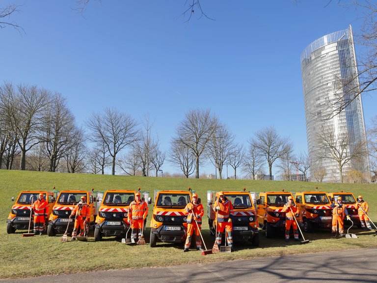 Acht Elektrofahrzeuge der bonnorange AöR mit ihren Fahrern, die Kehrbleche und Besen halten, stehen auf einer Wiese vor dem Posttower in Bonn. 