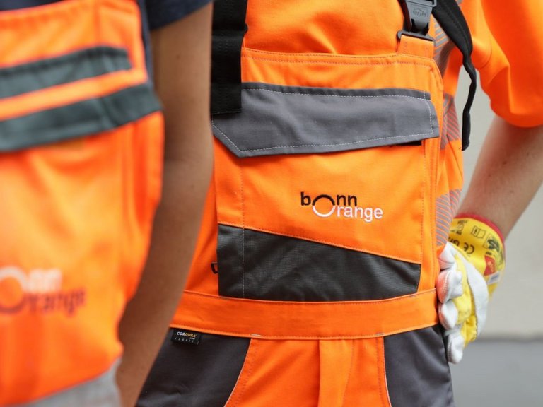 bonnorange gibt Änderungen bekannt Zwei Mitarbeitende der bonnorange AöR in Arbeitskleidung mit den Hände mit Arbeitshandschuhen in den Hüften, der Vordergrund ist unscharf.