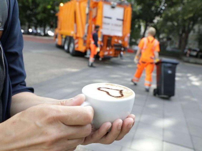 Tag der Daseinsvorsorge Foto von einer Hand, die einen Kaffee hält. Im Hintergrund Müllwerker von bonnorange bei der Arbeit.