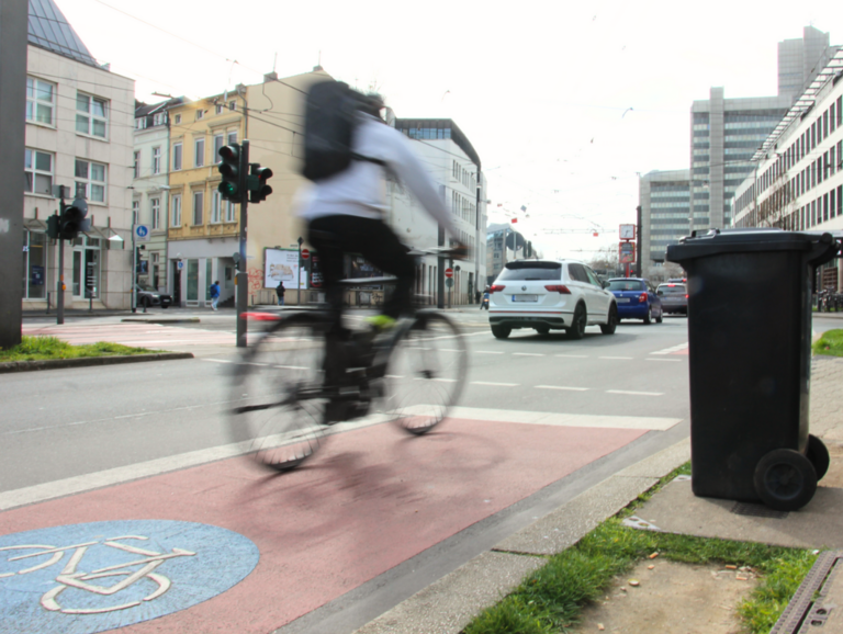 Bild einer Fahrradspur, auf der ein Fahrrad fährt. Daneben eine schwarze Mülltonne.